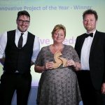 A woman in a formal dress stands in front of two other people in suits and holds a wooden triangular "Innovative Project of the Year 2017" award, presented to Revive Leeds.