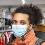 The image features a headshot photo of a person with black curly hair, in a orange jacket, wearing a face mask and looking at the camera