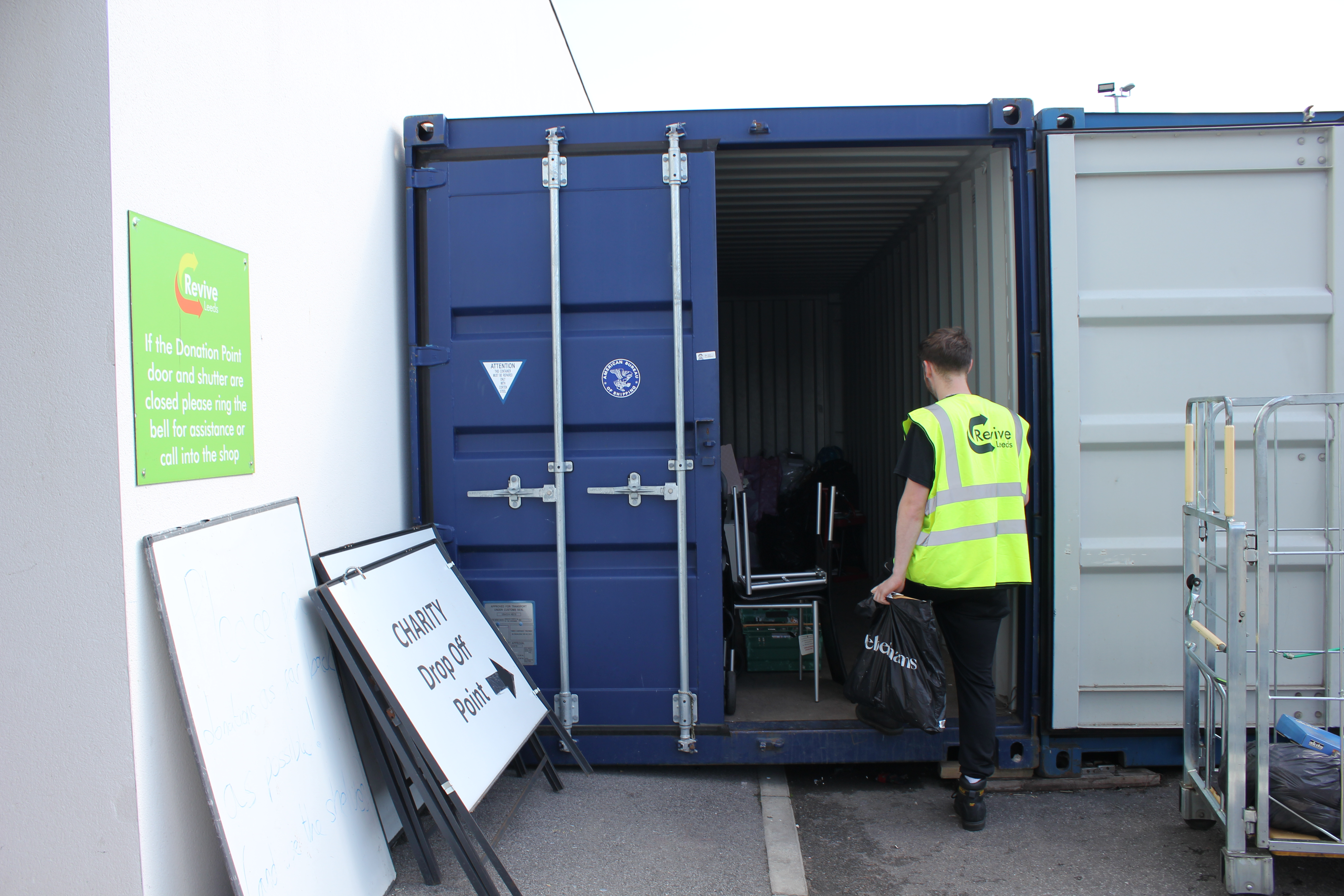An open blue freight container with a Revive Reuse staff member, wearing a high-vis jacket, stepping inside. A sign that reads 'Donation Point' is directed at the container.