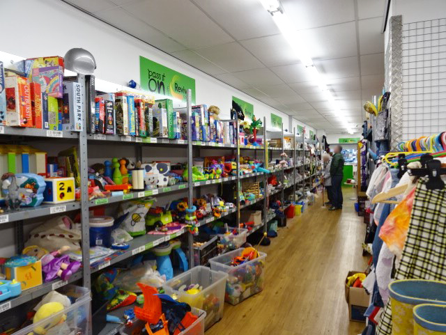 Revive Reuse Batley Shop 1.0 The image features a the inside of a Revive Reuse shop. On the left are metal shelves full of various kids toys. Two people are stood looking at the shelves. On the right is a metal clothes rail which is full of kids clothing items.