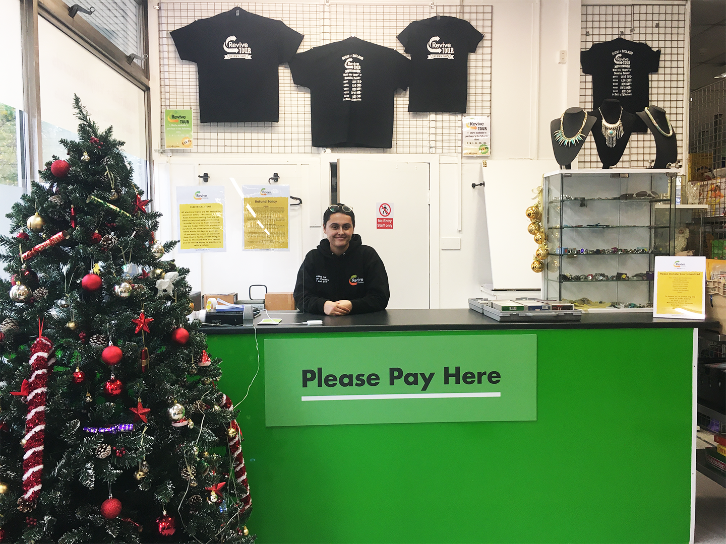 A Revive Reuse staff member working on the till behind the shop counter. The counter has a sign displayed that reads "Please Pay Here".