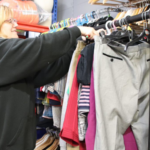 A female Revive Reuse staff member is hanging up various clothing items on a black clothes rail.