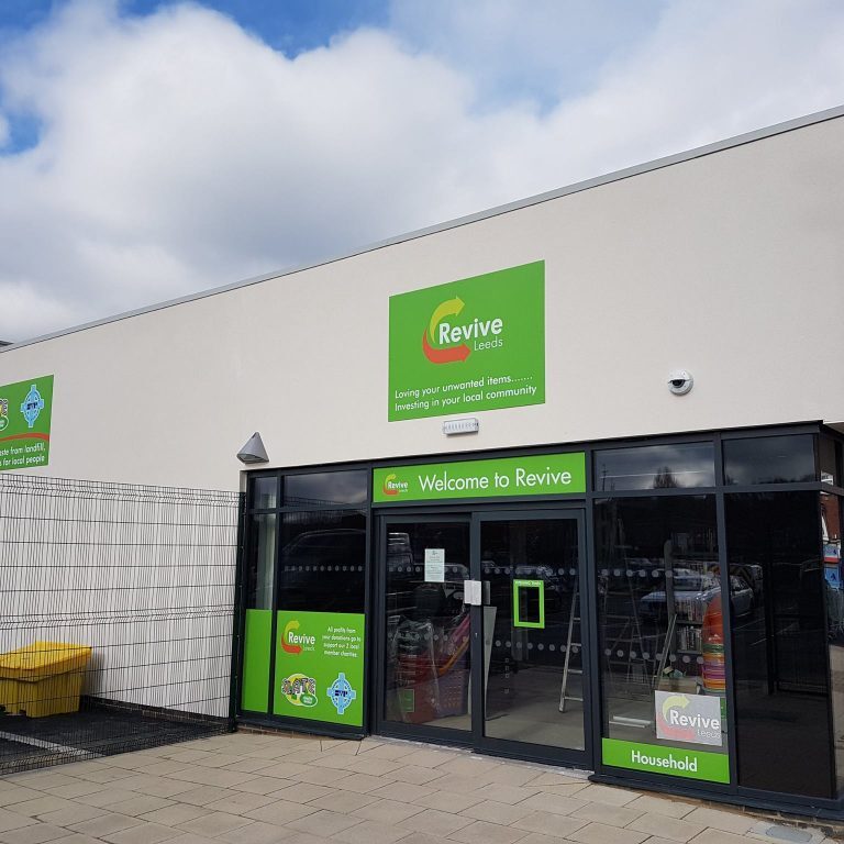 The image features a white building with many glass window and a green sign above the entrance that reads "Revive Leeds". "Loving your unwanted items...…. Investing in your local community". There is a car park to the left of the building.