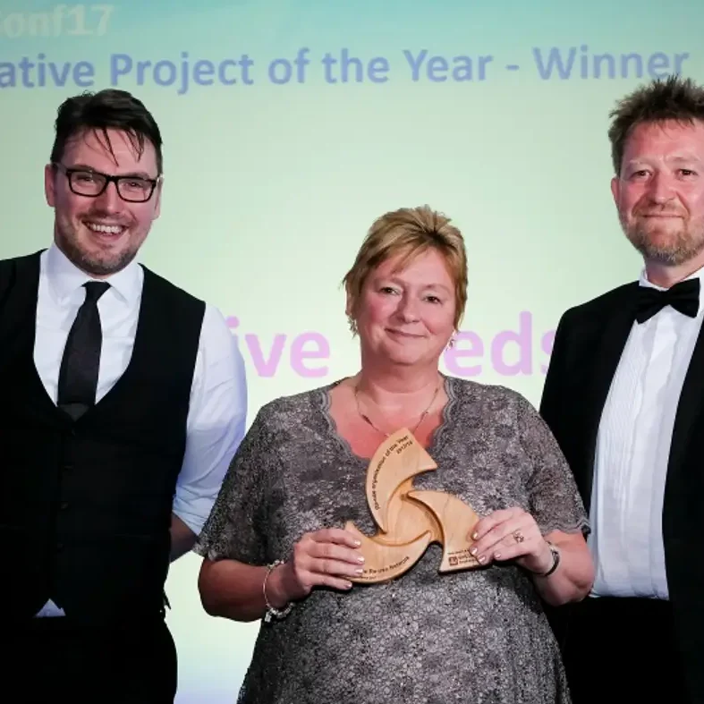 A woman stands in front of two other people in suits and holds a wooden triangular 'Innovative Project of the Year' award.